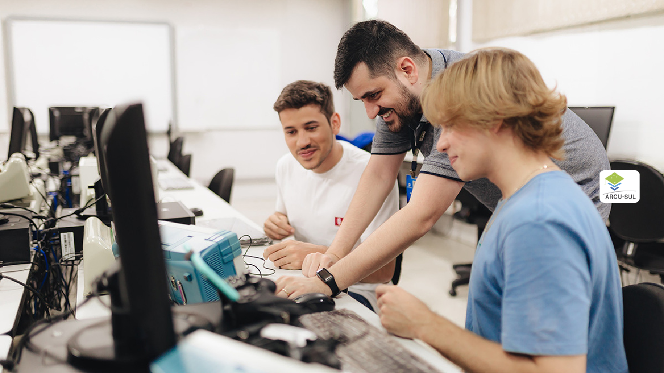 Professor ensina alunos enquanto manipula equipamento em meio a computadores